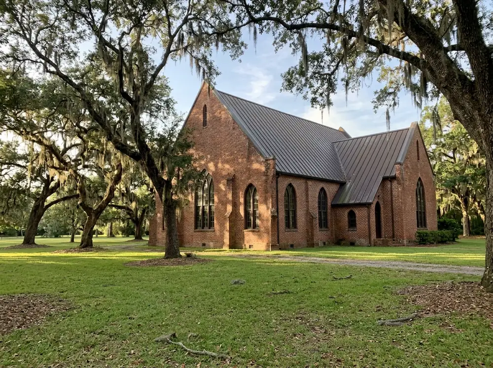 Steep standing seam metal roof in dark bronze on a brick sanctuary with tall gables, seen from a lawn with live oaks