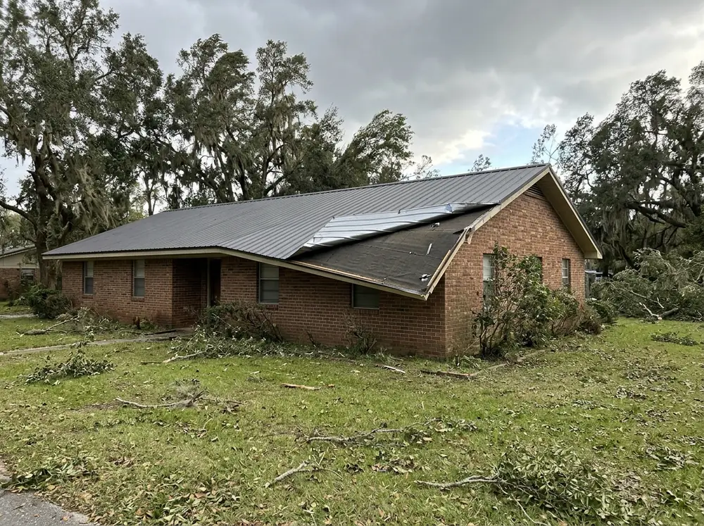 Gray metal roof with one panel edge peeled up at the rake and a section of trim missing after high wind