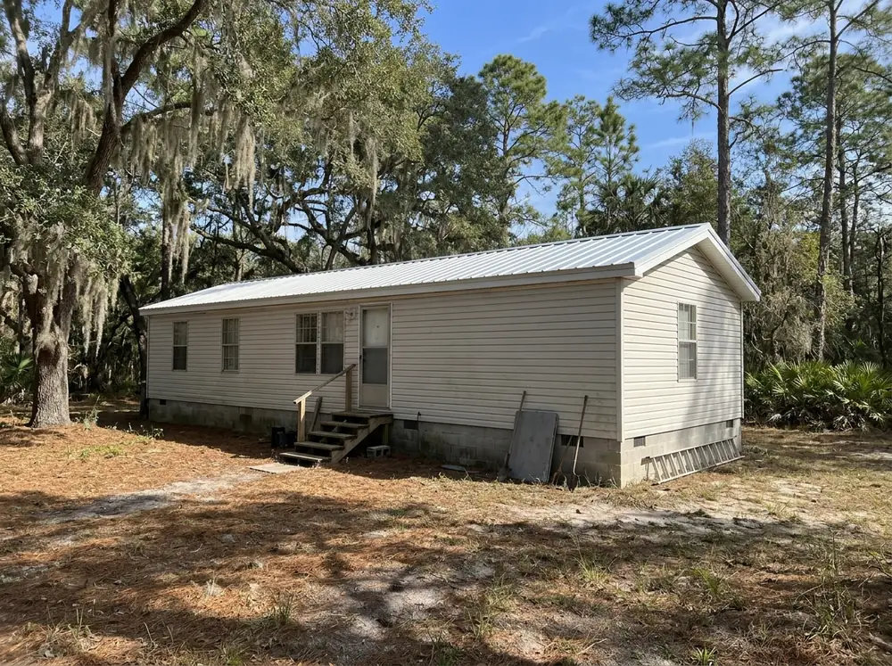 Manufactured home on a skirted block foundation with new white ribbed metal panels over the old roof, under live oaks